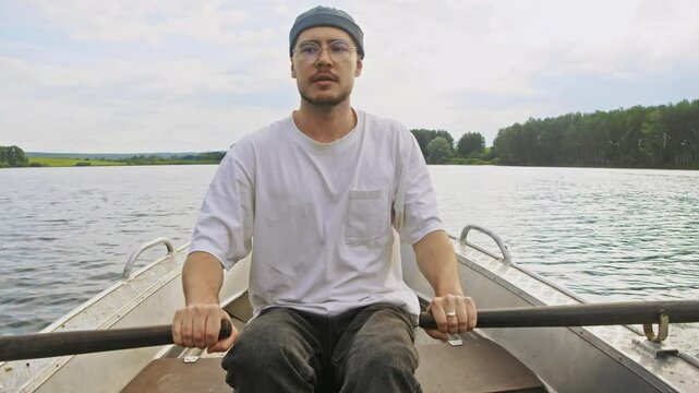 A young man with a beard and wearing a fishing hat is paddling a metal rowboat on the river on a summer's day. Portrait.