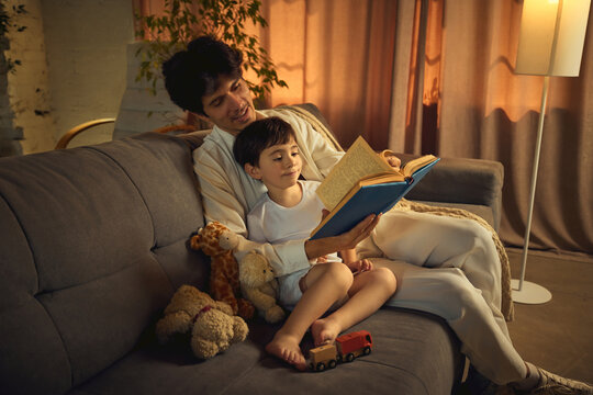 A father engages his son with a bedtime story while they sit on a comfortable sofa amidst warm lighting and scattered toys. Concept of parent-child bond and early childhood education - Powered by Adobe
