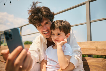 A father along with his young son capture a selfie while seated on a sun-drenched bench. Concept of...