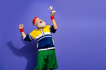Energetic senior man wearing retro sportswear joyfully holding a trophy in celebration against a...