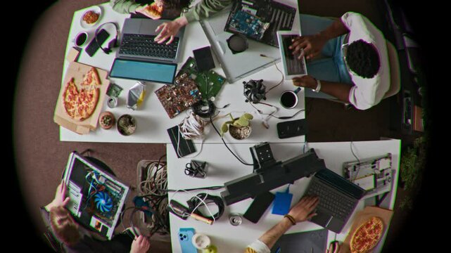 Fish eye directly above shot of cluttered tables with young people working on laptops, surrounded by wires, circuit boards, coffee cups and pizza boxes in busy and collaborative tech atmosphere