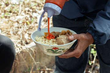 Hands squeezing chili sauce onto a bowl of instant noodles and meatballs outdoors
