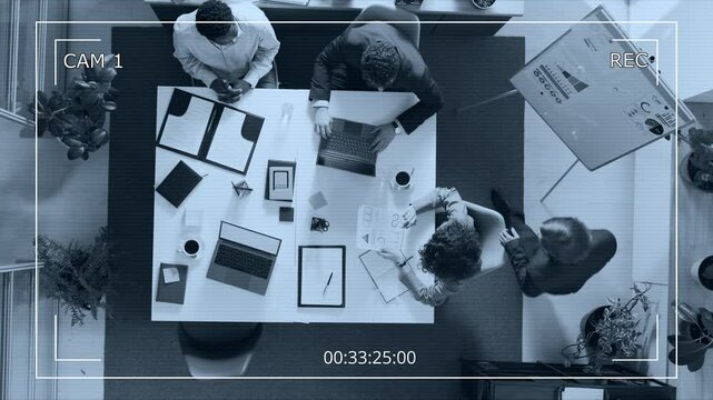 High angle surveillance camera shot of man coming up to woman sitting at white table and they analysing documents together in office setting with two other managers