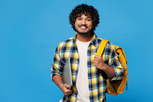 Young smiling smart IT Indian boy student wear yellow casual clothes backpack bag hold closed laptop pc computer look camera isolated on plain blue background. High school university college concept.