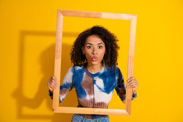Young woman holding wooden frame playfully on vibrant yellow backdrop wearing tie-dye shirt with...