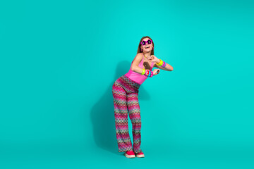 Young and trendy woman in vibrant retro outfit posing against a solid turquoise background,...