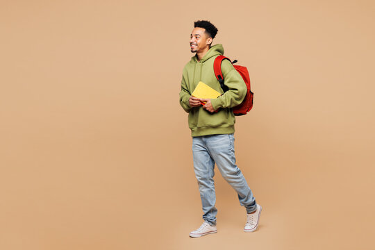 Full body side view young smart boy student wearing green casual clothes backpack bag hold books walking going isolated on plain pastel light beige background. High school university college concept.
