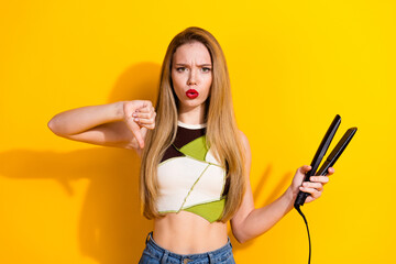 Upset young woman holding a flat iron showing a disapproval thumbs-down gesture symbolizing...
