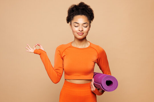 Young fitness trainer instructor sporty woman wear orange clothes spend time in home gym hold purple yoga mat do om gesture meditate isolated on plain beige background. Workout sport fit abs concept. - Powered by Adobe