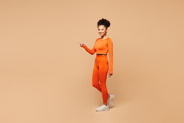 Full body side view young fitness trainer instructor sporty woman wear orange clothes spend time in gym walk go point aside on area isolated on plain beige background. Workout sport fit abs concept. © ViDi Studio
