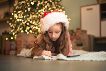 little girl in santa hat lying on floor at home writing Christmas wish list, letter to Santa, dream...