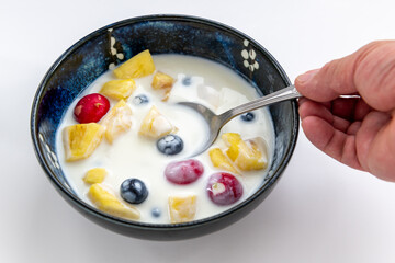 A person using a spoon to eat a bowl of mixed fruit and kefir natural yogurt, isolated on a white background.