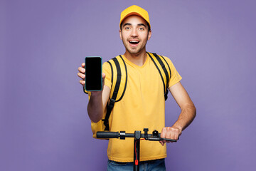 Young delivery guy man he wearing yellow cap t-shirt uniform workwear thermal bag work as dealer courier ride e-scooter show blank screen mobile cell phone isolated on plain pastel purple background.