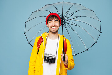 Traveler man he wear hat yellow raincoat casual clothes hold umbrella isolated on plain pastel blue background. Tourist travel abroad in free spare time rest getaway. Air flight trip journey concept.