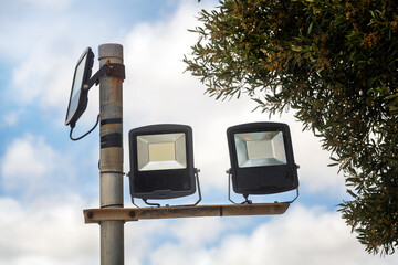 Modern LED floodlights mounted on a metal pole, with a solar panel above and green tree branches on...