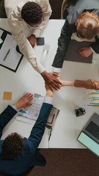 Vertical top down shot of multiethnic team of three young men and unknown woman stacking hands in centre of office desk