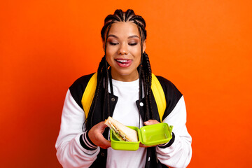 Teenage girl holding a sandwich in a green lunchbox while smiling against an orange background...