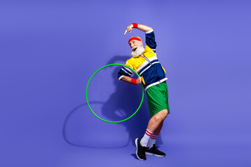 Senior athlete in retro athletic outfit demonstrating fitness with green hula hoop against purple...