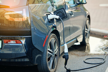 Vehicle connected to electric charging station at urban parking lot after rainfall with wet cable, posing potential safety risks from electricity. Electric vehicle charging at public station