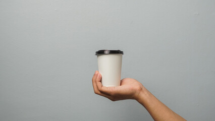 Hand holding white disposable coffee cup with black lid on grey background for branding mockup.