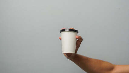 Hand Holding White Disposable Coffee Cup with Black Lid Against Plain Grey Background