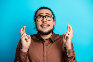 Young man in brown shirt crossing fingers and looking hopeful against turquoise background