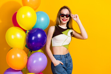 Young woman in stylish casual outfit with balloons and vibrant yellow backdrop, exuding happiness...