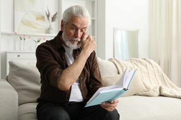 Senior man with vision problem reading book on sofa at home, space for text