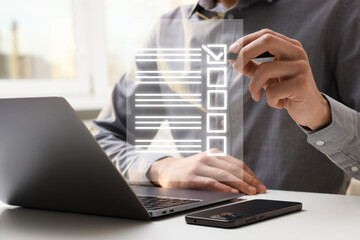 Man putting tick in check list on virtual screen at table with laptop and mobile phone, closeup