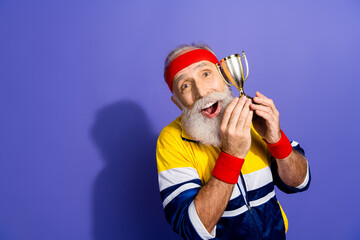 Cheerful elderly man in retro athletic wear celebrates achievement with small gold trophy standing...
