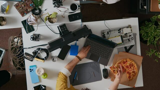 Directly above shot of young man wearing yellow shirt using laptop with one hand and holding slice of pepperoni pizza in other, sitting at cluttered white desk filled with electronic components