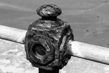 Close-up, black and white view of a rusty, weathered coastal railing post with a decorative top