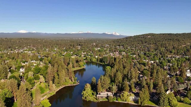 Bend, Oregon, United States. Aerial view of Mirror Pond reflecting the crisp blue sky amidst the town's vibrant green trees, with snow-capped three sisters mountains in the distance