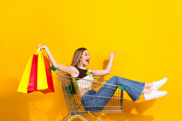 Young woman shopping joyfully, seated in shopping cart while holding bags against a yellow...