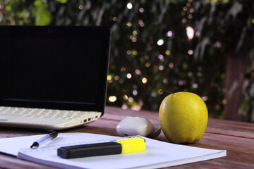 A laptop lying on a wooden surface outdoors. Next to the laptop is a yellow apple, as well as an open notebook with a pen and a yellow marker. 
