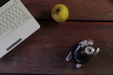 There is a microscope, a laptop and a green apple on the wooden table.