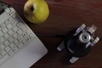 There is a microscope and a laptop on the wooden table. The emphasis is on the apple.