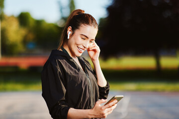 Sportswoman listening music on wireless earbuds and holding smartphone