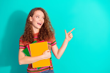 Smiling young student pointing to side holding notebooks against a teal background showcasing...