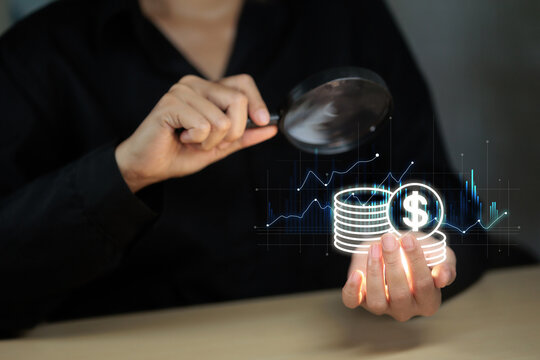 A person uses a magnifying glass to inspect a holographic image of coins and a financial graph, symbolizing financial analysis and investment review.
