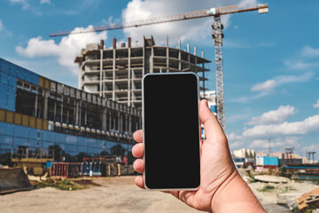 A hand holds a smartphone with a blank screen in front of a construction site with a crane. Perfect for tech and building industry content