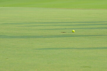 A golf ball near the hole on a green. The morning sun casts long shadows on the grass.