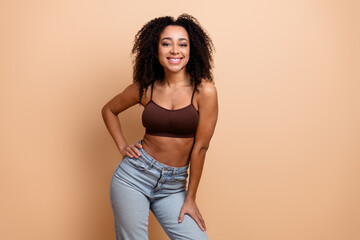 Young woman in casual brown top and jeans smiling against beige background exuding confidence and...