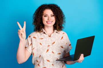 Smiling young woman holding a laptop while making a peace gesture against a bright blue background