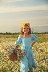 Natural happy girl in blue linen dress and yellow knitted kerchief stands and holds bouquet of flowers in daisy field