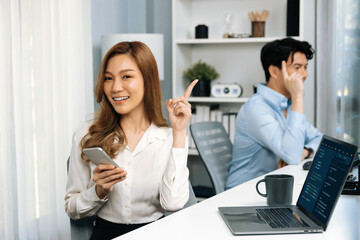 Profile smiling beautiful Asian businesswoman looking camera, using phone for advertisement at modern office on working desk casual day. Blurry background man colleague analyzing data on pc. Infobahn.