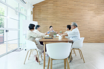 Group of Southeast Asian elderly friends sitting around wooden table in bright modern living room, sharing tea and conversation in warm, natural light, symbolizing friendship and happy retirement life