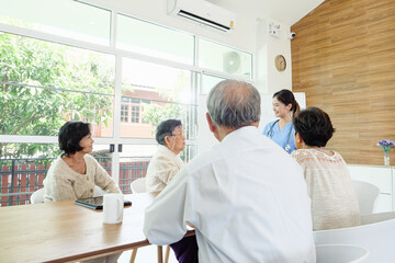 Asian healthcare professional in blue scrubs leading discussion with group of senior patients in bright modern clinic, explaining medical care plans and promoting healthy lifestyle awareness.
