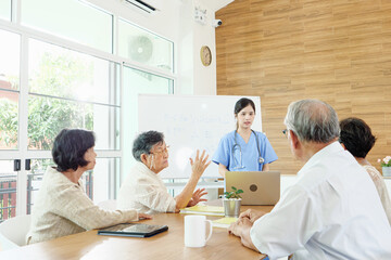 Asian healthcare professional in blue scrubs leading discussion with group of senior patients in bright modern clinic, explaining medical care plans and promoting healthy lifestyle awareness.
