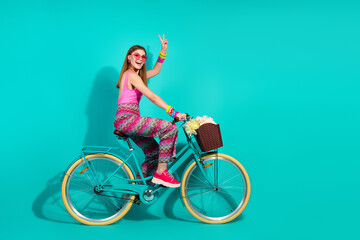 Young smiling woman on a teal bicycle with flowers in basket on vibrant turquoise background in...